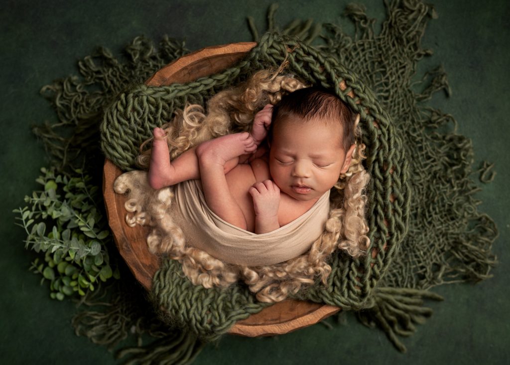 baby boy curled up in a wooden bowl with green foliage during a newborn photoshoot in kent