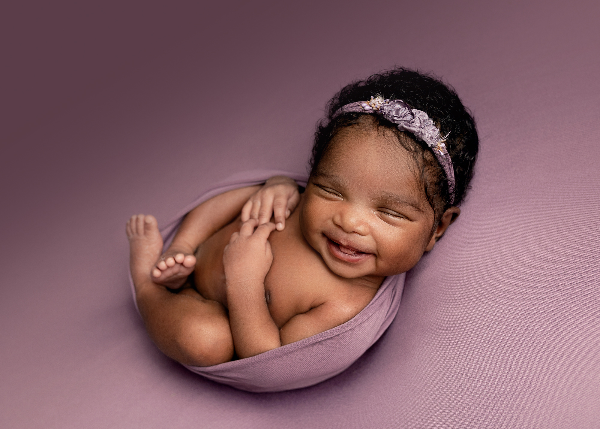 newborn wrapped in purple blanket smiling at camera being photographed by photographer in Kent
