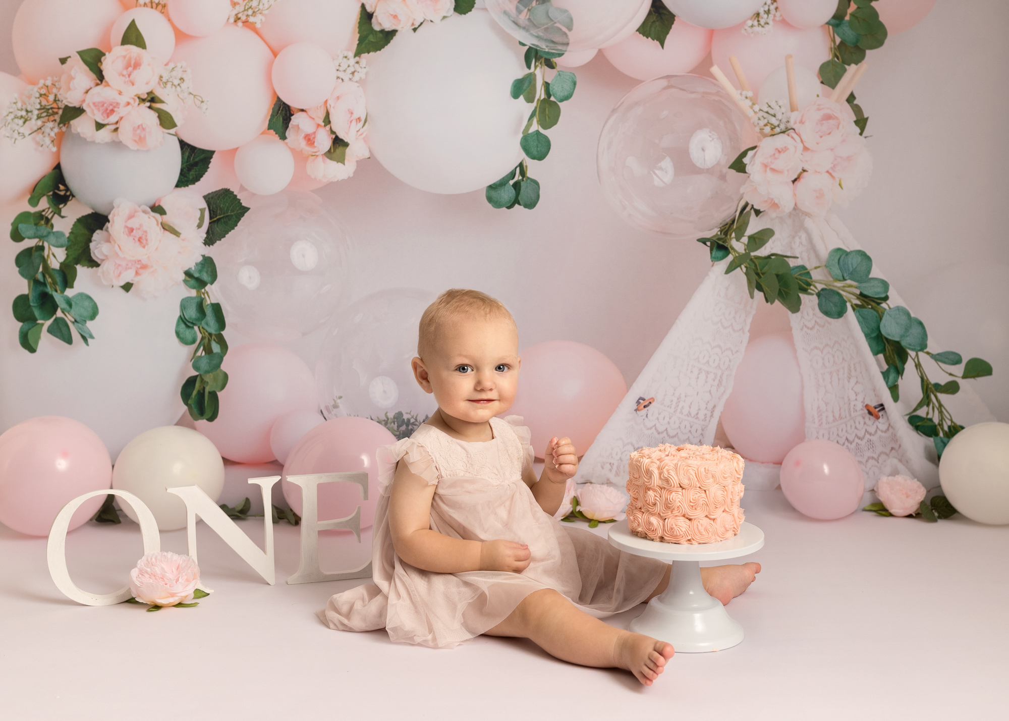 Baby girl eating cake off a wooden spoon sat next to a rainbow cake and rainbow balloon arch during cake smash photoshoot in Kent