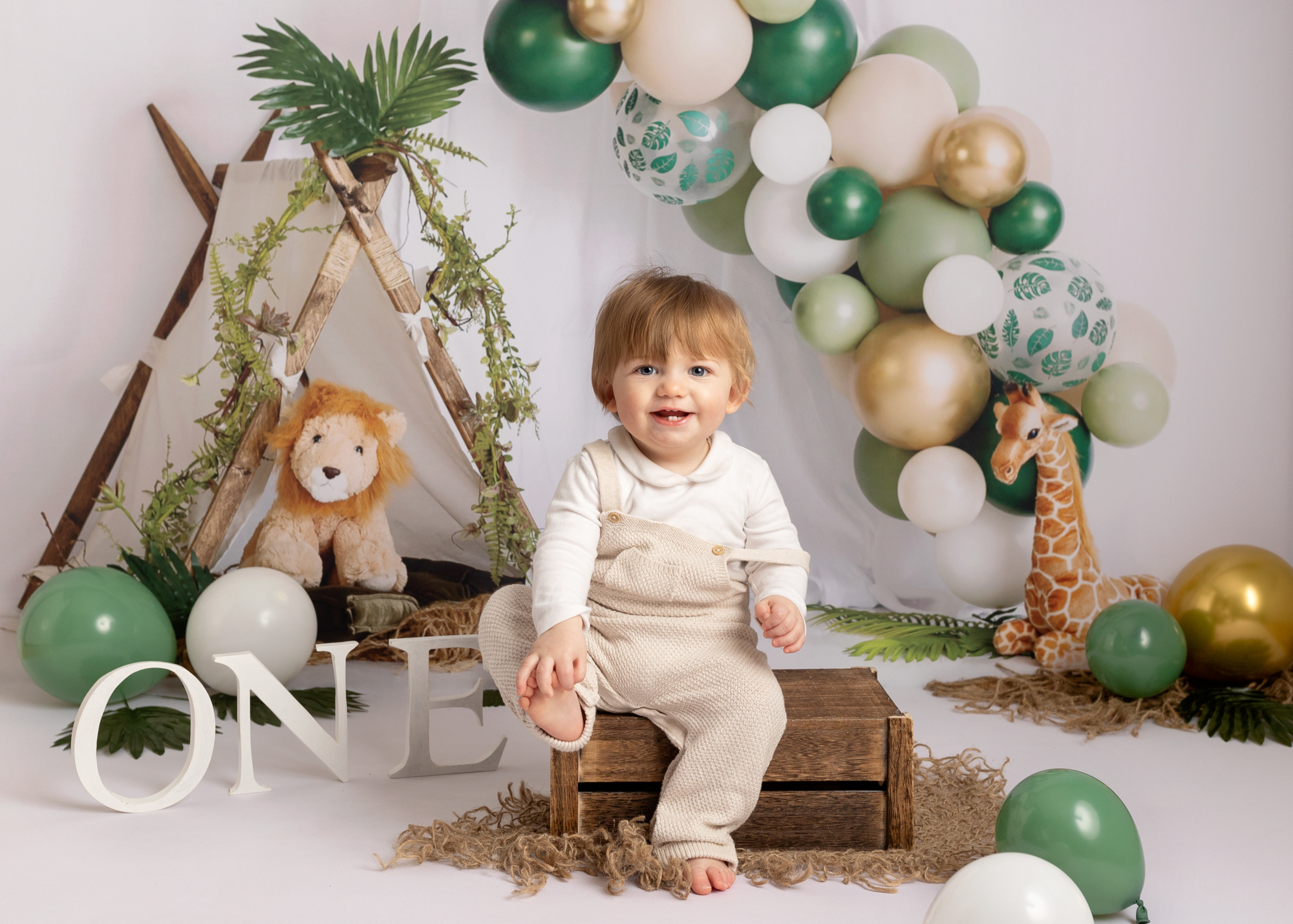 Boy sitting on wooden box with jungle theme backdrop hiding his foot and smiling at the camera, during a birthday photoshoot in Kent