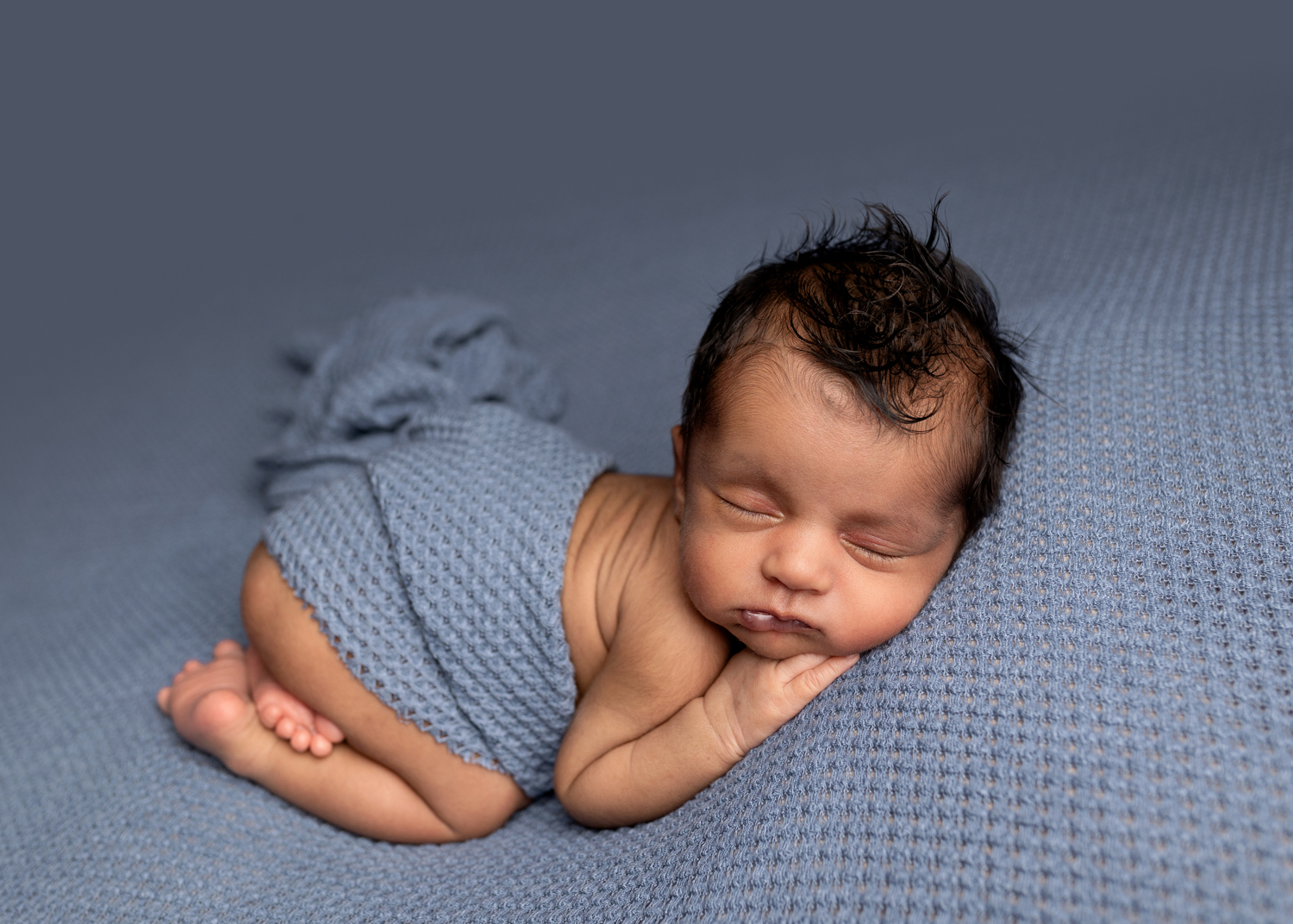 Baby boy curled up on a blue blanket with jet black hair, photographed in Kent