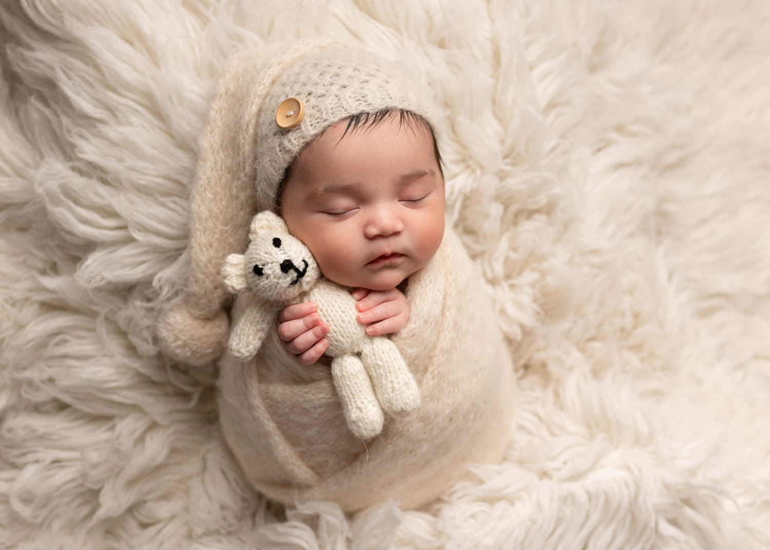 newborn baby girl wrapped up in cream wrap, cuddling a teddy on a white fluffy rug. Photographed during a newborn photoshoot in Kent