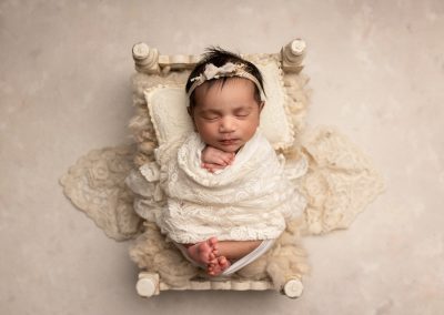 newborn baby girl wrapped in cream lace, laying in a white wooden bed with a cream headband during a newborn photoshoot in Kent
