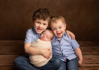2 boys holding their baby brother wrapped in white blanket and smiling for a photoshoot in Kent
